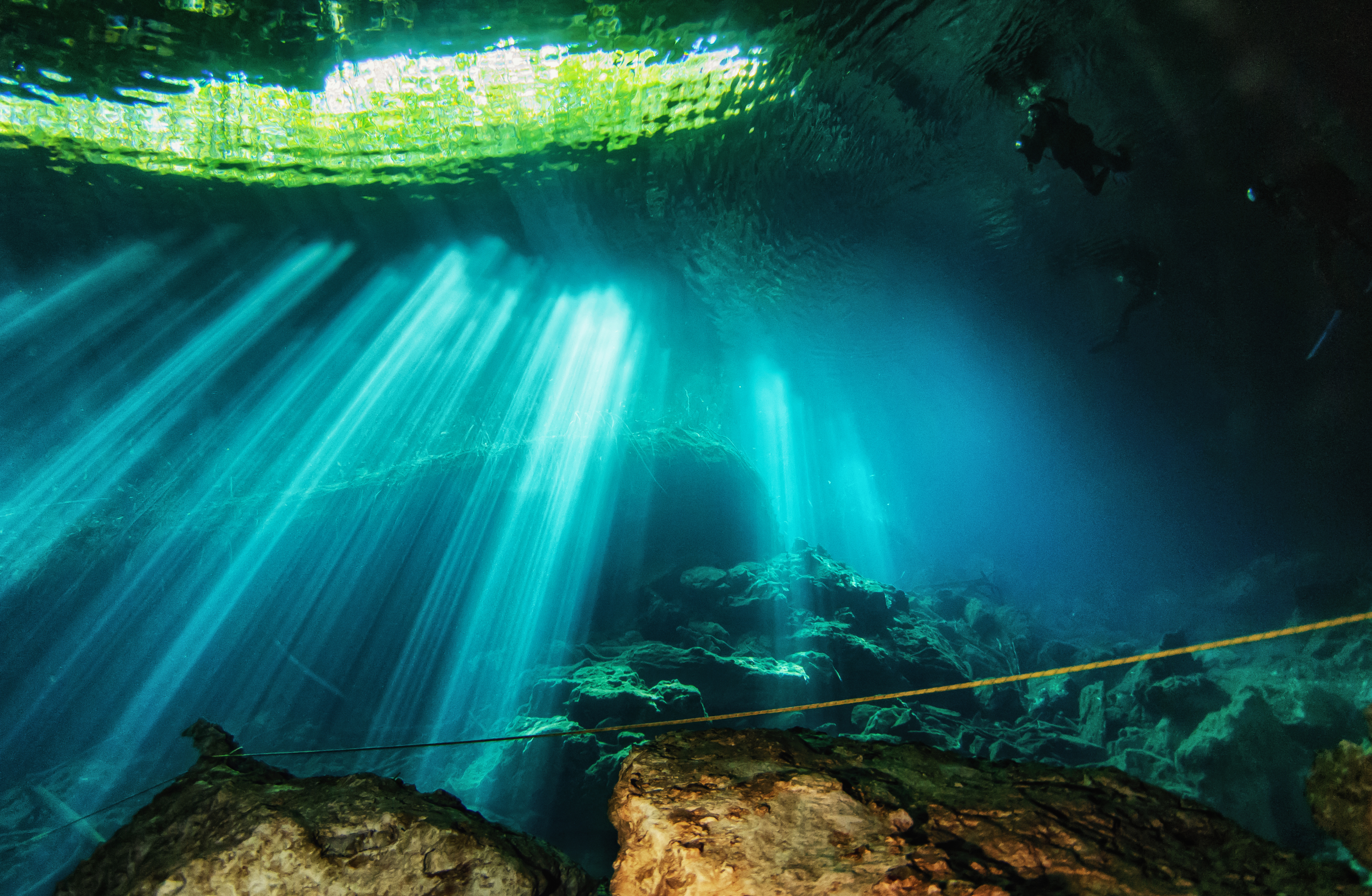 Scuba divers explore a cenote in Playa del Carmen, Mexico as sunlight streams through the water, illuminating the rocky underwater scene.