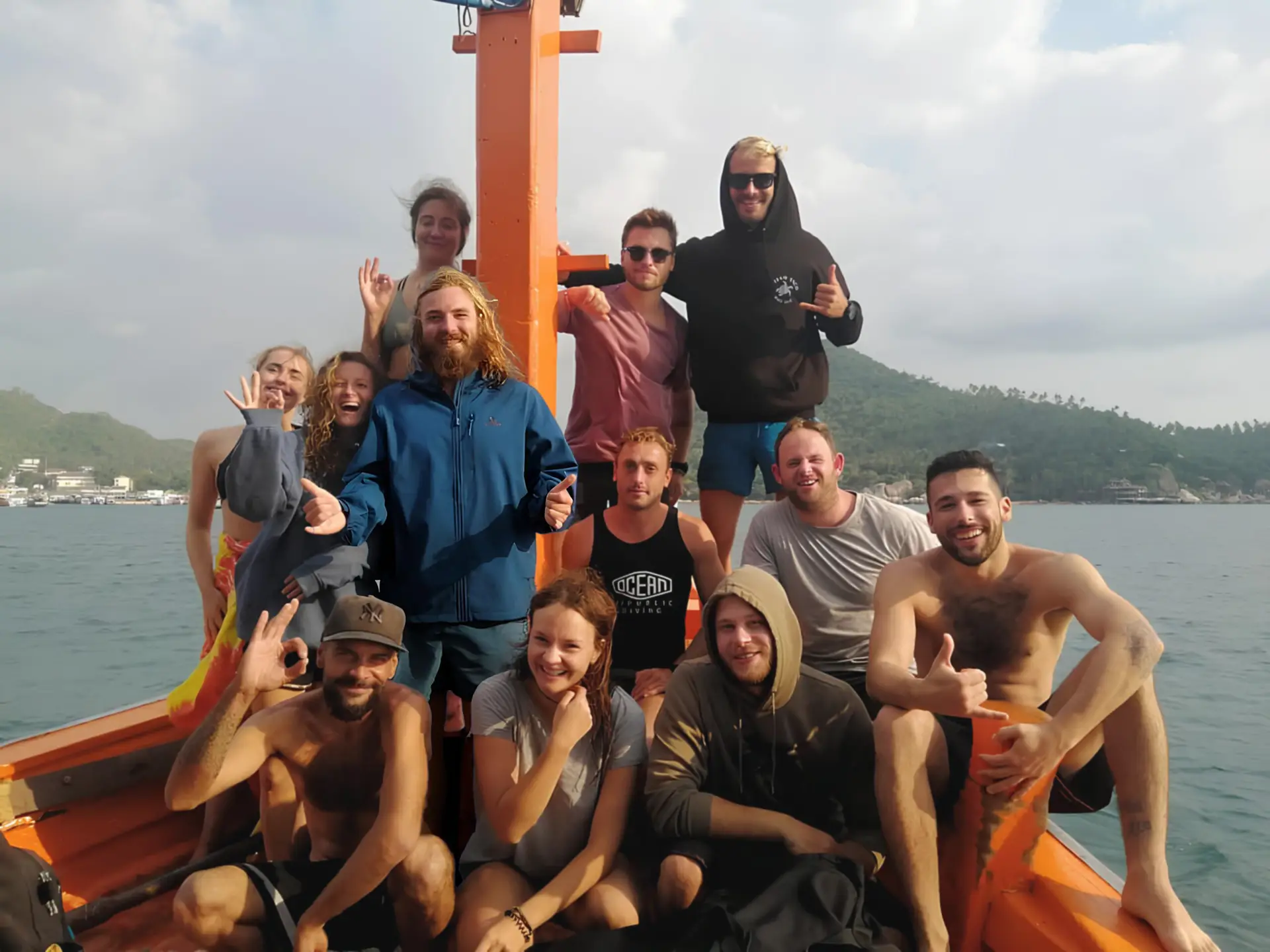 Group of 11 scuba diving students and instructors on a boat after a PADI Open Water course, with mountains and sea in the background, some giving thumbs up and dive hand signs