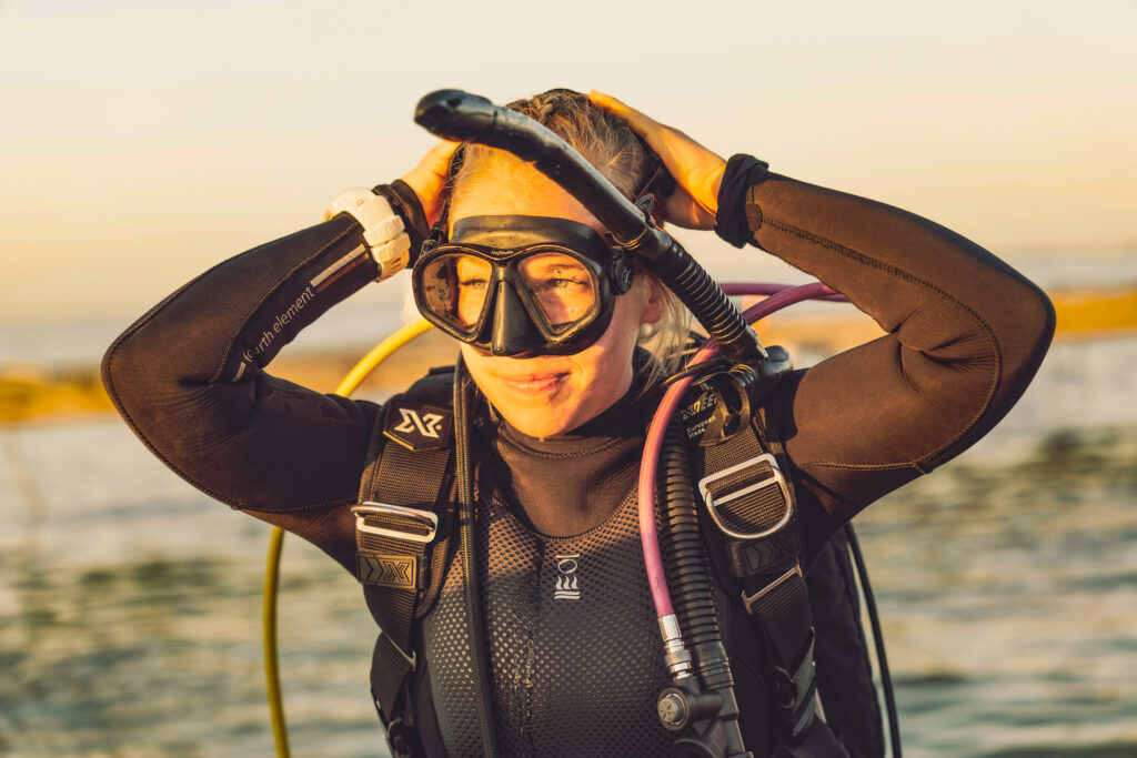 Diver in a black wetsuit adjusting her mask at the water's surface during golden hour.