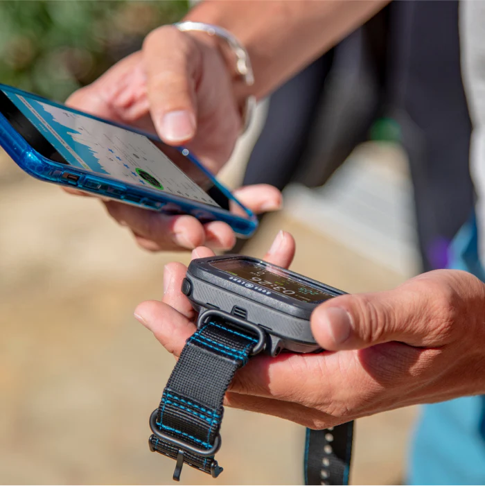 Close-up of hands holding a dive computer and a smartphone outdoors, syncing or reviewing dive data.